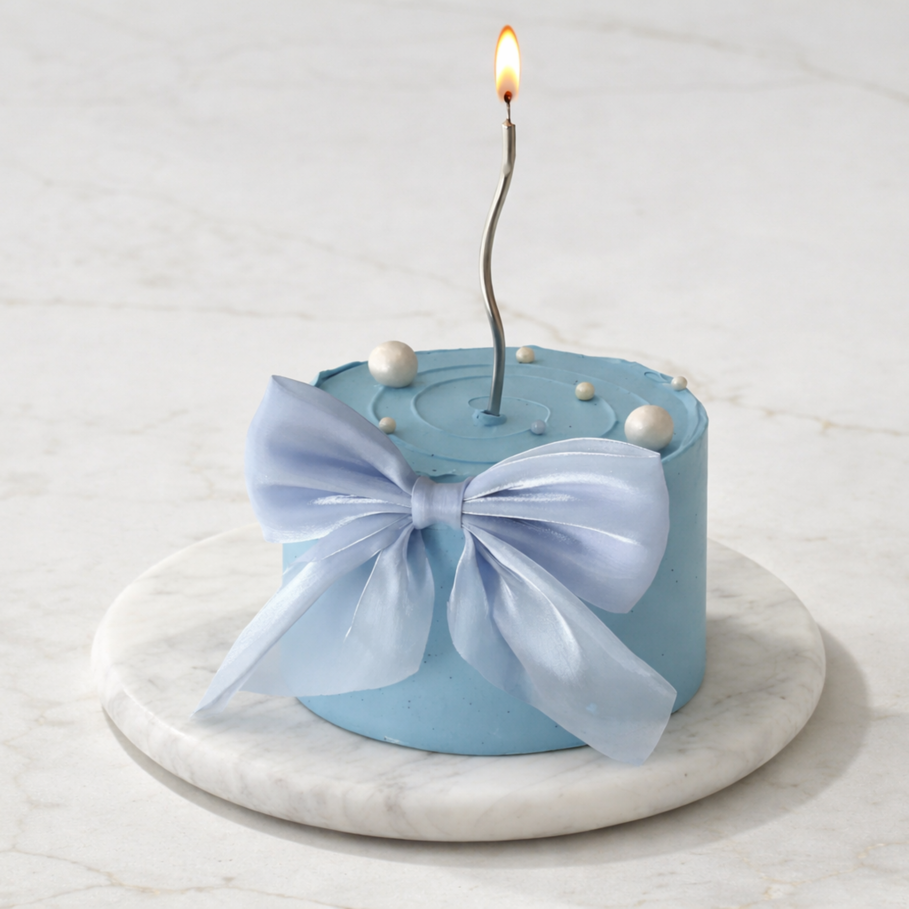 Blue cake with a white bow and lit candle on a marble stand against a light background