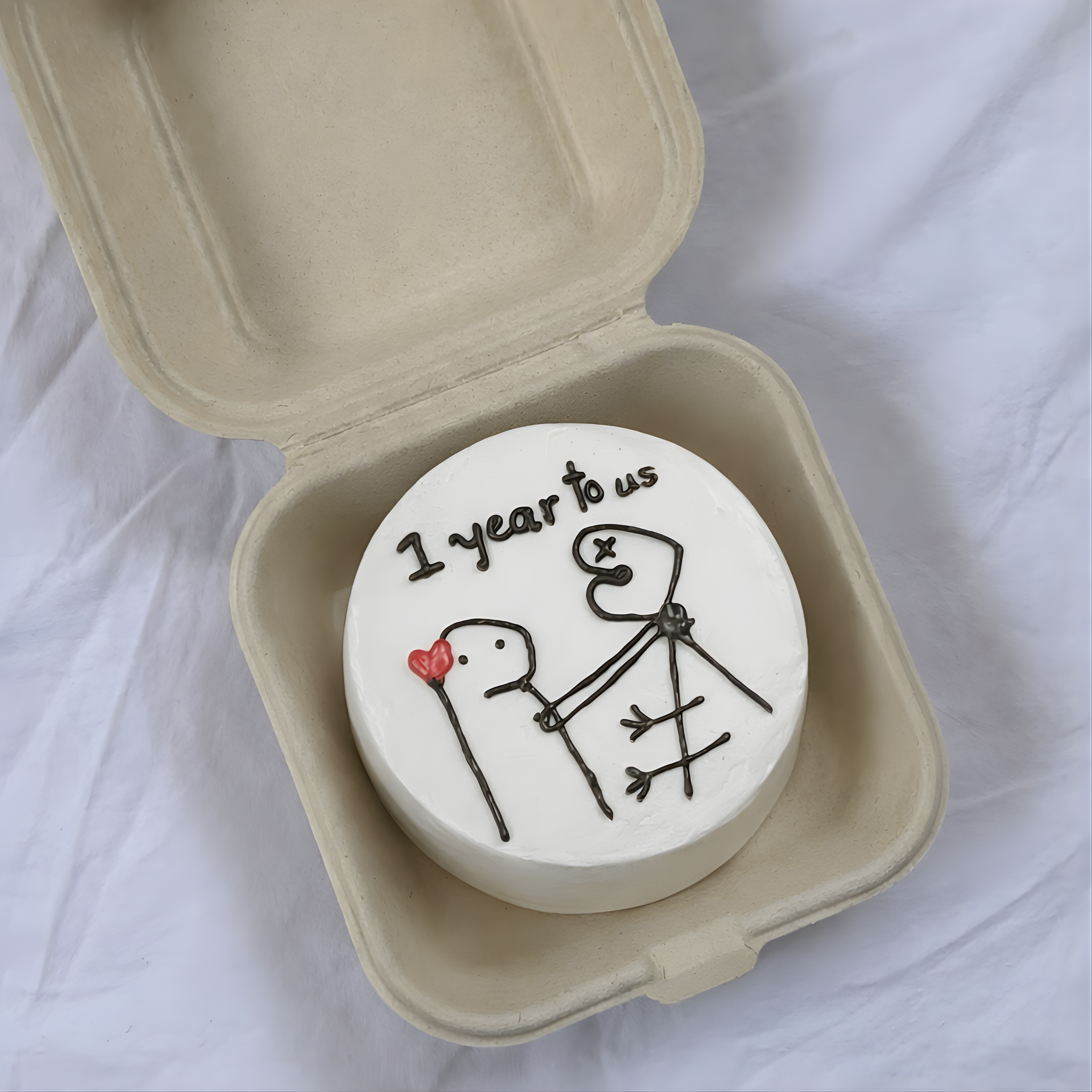 Small round cake with '1 year to us' text and stick figure design in a cardboard container.