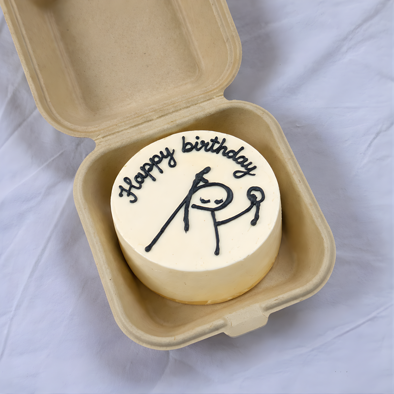 Small round birthday cake with 'Happy Birthday' and stick figure design in a cardboard container on a white background