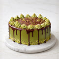 Rich pistachio chocolate cake with ganache drips, creamy frosting, and pistachio topping on a marble platter.